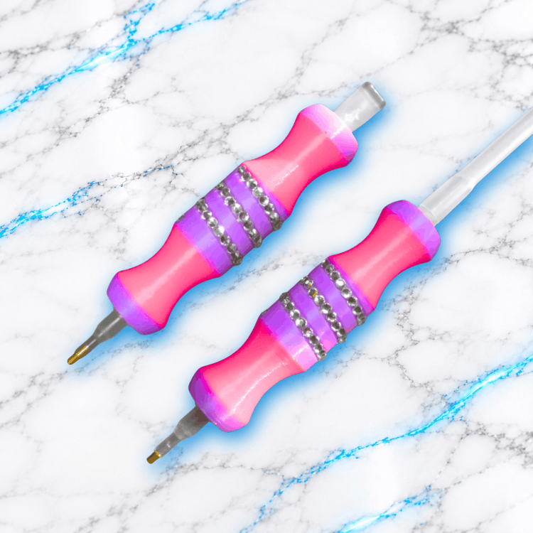 Deux crayons Diamond Painting Medium Barbu Glitter roses et violets ornés de strass argentés, l’un avec embout manuel et l’autre relié à un tuyau d’air.