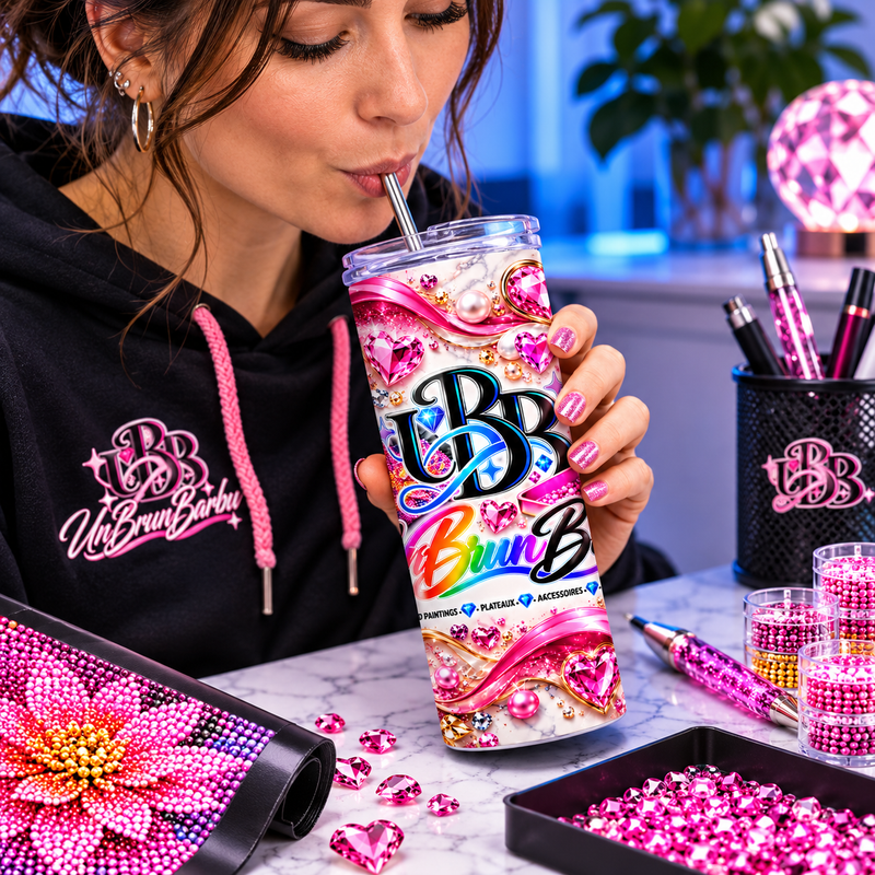 Femme buvant à la paille dans le gobelet isotherme UBB Éclat Rose, au milieu d’une toile fleurie et d’accessoires de Diamond Painting.