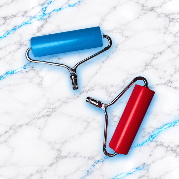 Deux Roller Press pour Diamond Painting UnBrunBarbu, modèles bleu et rouge avec supports métalliques, rouleaux de pression pour fixer les diamants sur une toile de broderie diamant, sur fond marbre blanc veiné de bleu électrique.