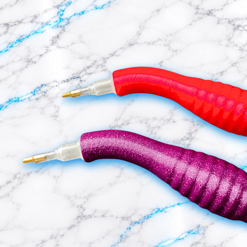 Gros plan sur les embouts de deux stylos ergonomiques courbés pour broderie diamant, avec pointes métalliques et manches 3D rouge et violet.