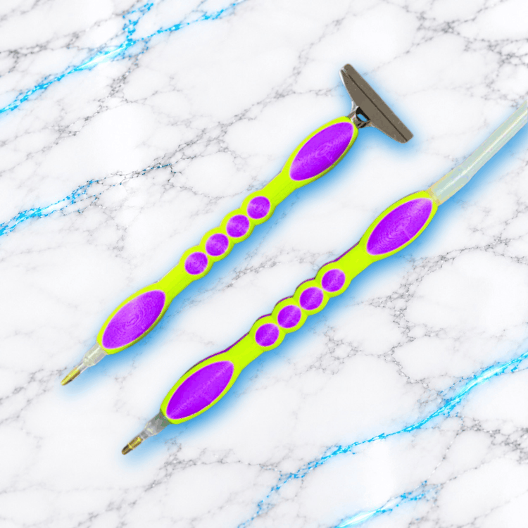 Deux crayons Diamond Painting Dessous Plat Classique vert fluo et violet, l’un avec embout large métallique et l’autre relié à un tuyau d’air transparent.