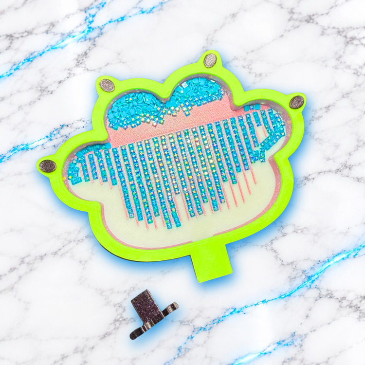 Plateau Diamond Painting en forme de patte de chat à bordure vert fluo, avec diamants turquoise alignés, rainures roses et bouchon pailleté retiré devant.